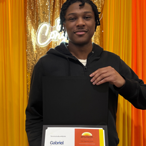 A person in a black sweatshirt holds an orange and yellow certificate in front of an orange and yellow curtain backdrop, with a partially obscured white neon sign behind them.