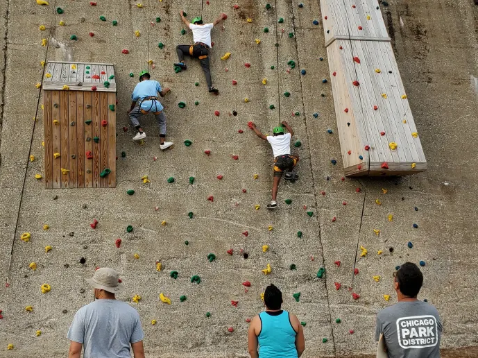 Six people at an indoor rock climbing wall; three climb the light brown wall with colorful holds while three others watch from the foreground.