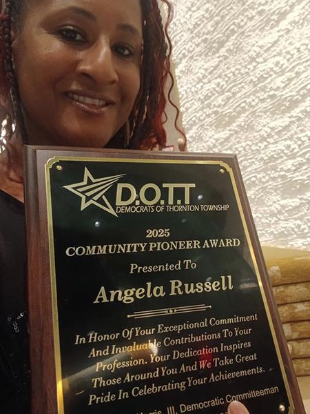 A person smiles at the camera while holding an award reading '2025 Community Pioneer Award Presented to Angela Russell.'