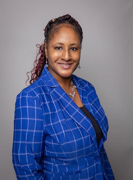 A person smiles at the camera in a portrait, wearing earrings, a necklace, and a blue plaid blazer.