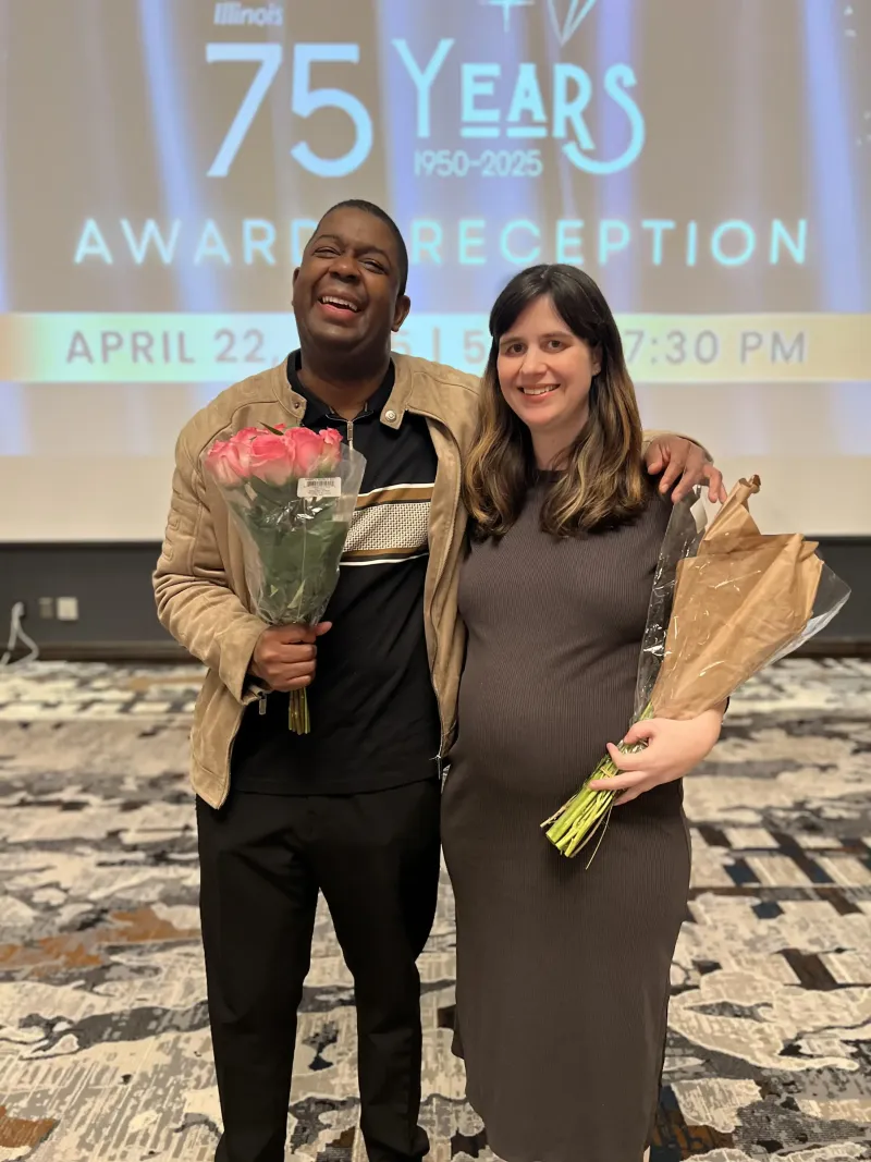 Two people smiling, holding bouquets, with a '75 Years Award Reception' screen in the background.