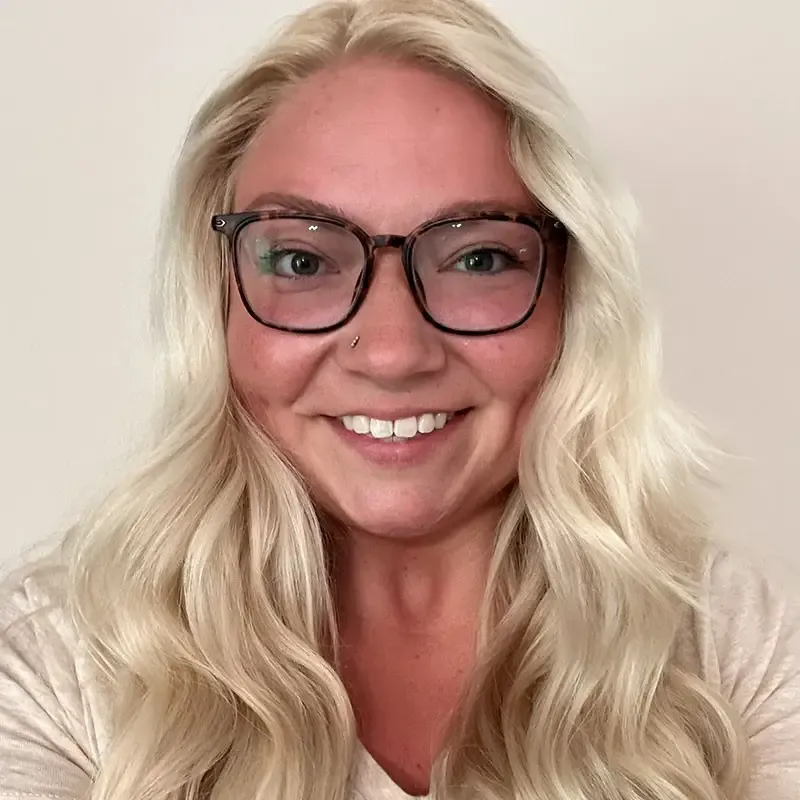 Person with long blonde hair and black-framed glasses smiling at the camera against a neutral background.