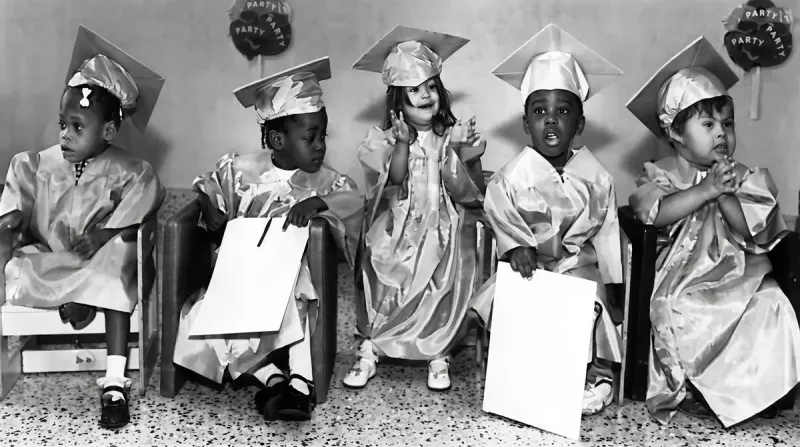 Five young children in graduation gowns and caps seated in a row, with 'PARTY' balloons in the background.