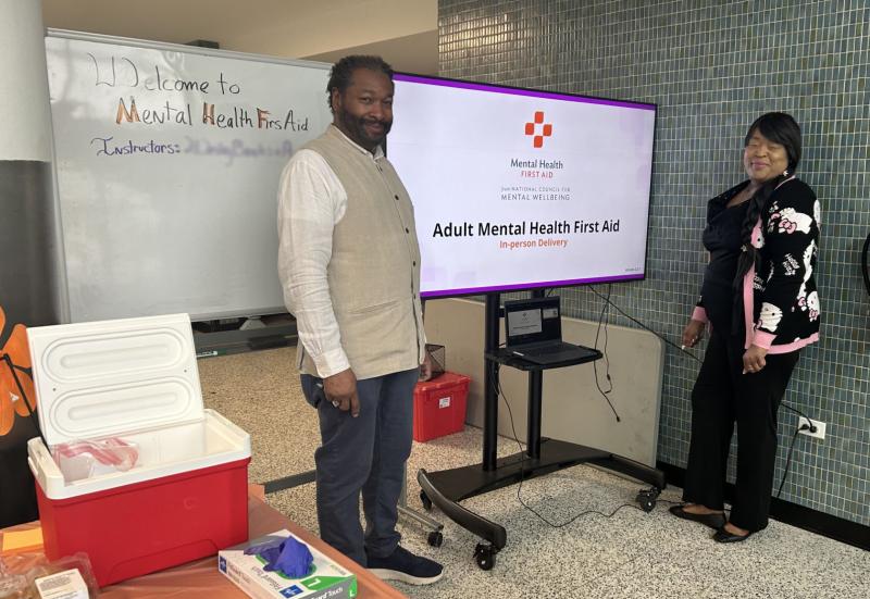 Two instructors stand in front of display materials regarding Mental Health First Aid training