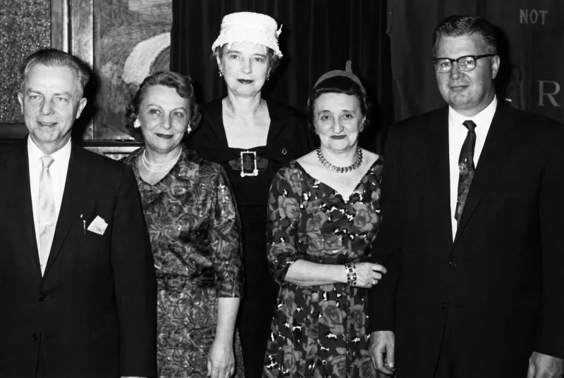 Five people pose in semi-formal attire, with three women in dresses and two men in suits.