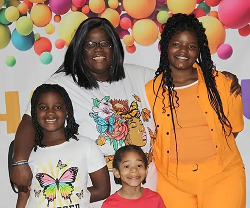 Four people smiling at a celebration or luncheon event, posed in front of a colorful backdrop of hanging balloons in various bright colors.