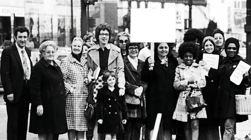 A black and white photo of a group of people at a rally, holding signs and smiling.