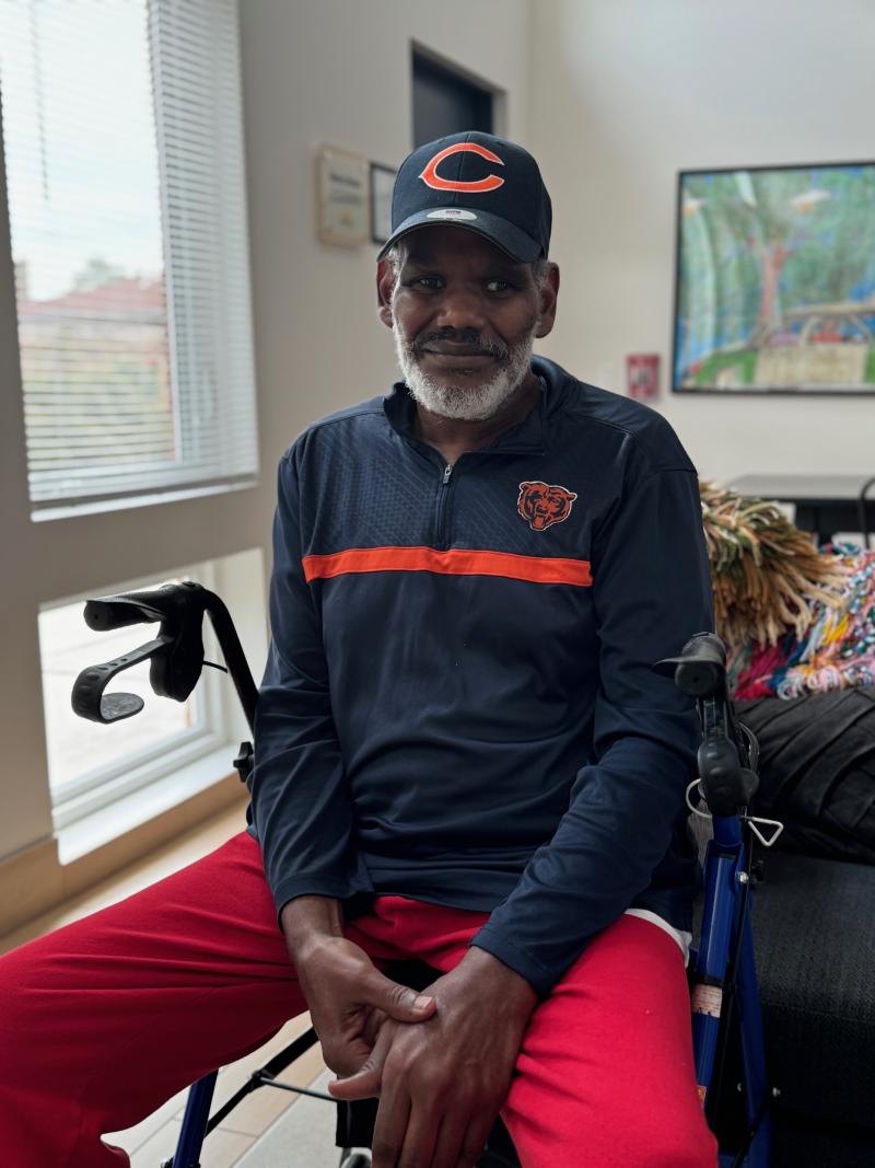 Man sitting on a mobility aid wearing a blue cap and jacket with red pants, next to a window.