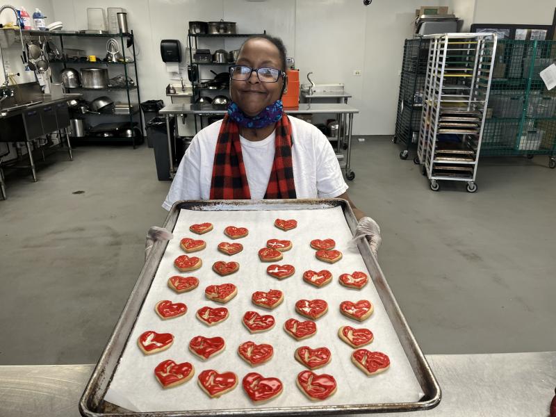 A person in glasses and a red and black plaid apron holds a tray of heart-shaped cookies with red icing in a kitchen.