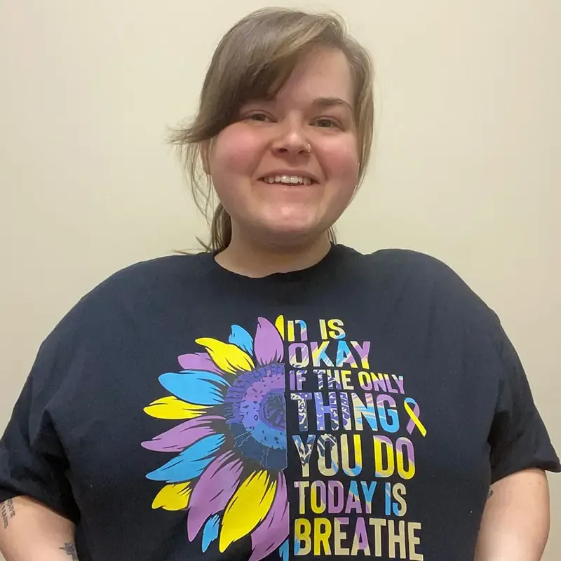 Person smiling at the camera wearing a dark t-shirt with a colorful flower graphic and motivational text about resilience and mental health support.