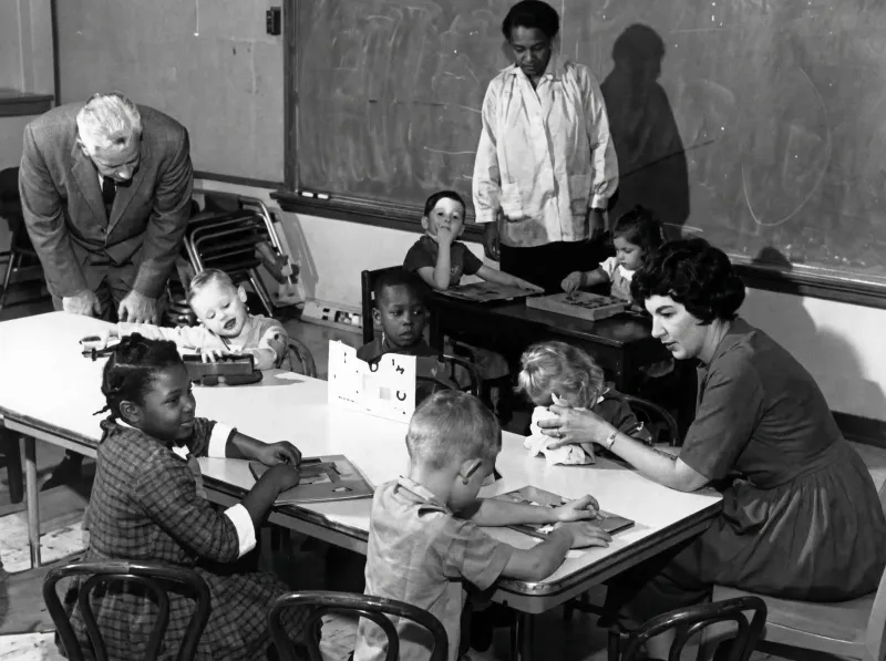 Children and adults in a mid-20th century classroom working together at tables with educational activities.