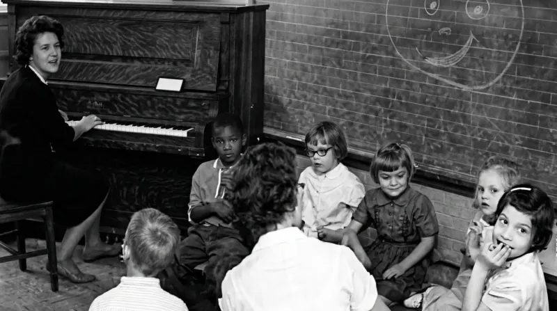 A woman plays the piano in a classroom with a group of children seated on the floor in front of her. A smiley face is drawn on the chalkboard behind them.