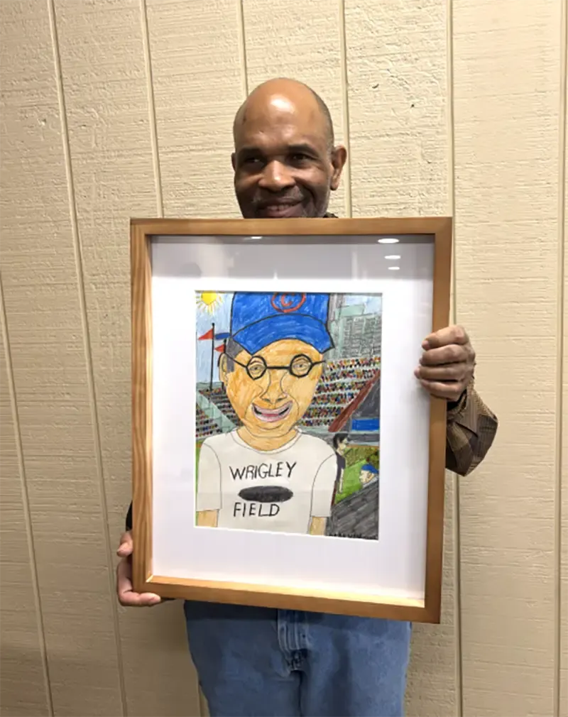 A person holds a frame with a colorful drawing of a smiling figure wearing a blue cap and glasses, with 'WRIGLEY FIELD' on their shirt.