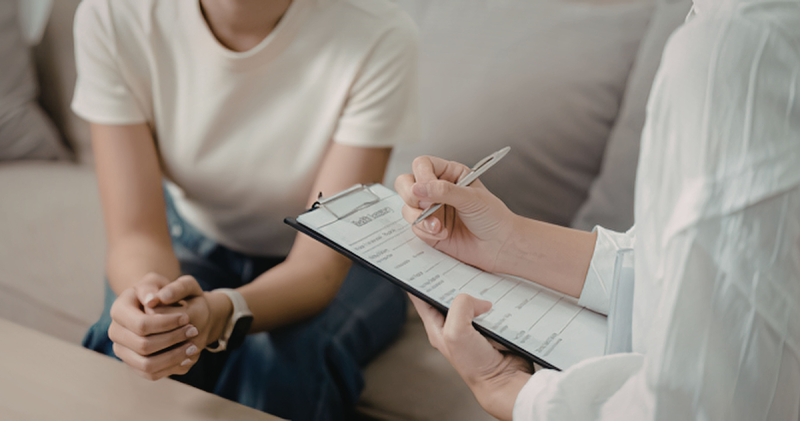 A person writes on a clipboard in the foreground while another sits on a couch leaning forward in the background.