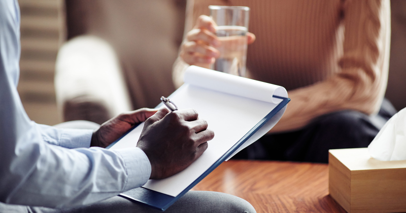 Two people meet across a table; one takes notes on a clipboard, the other holds a glass of water, with a tissue box between them.