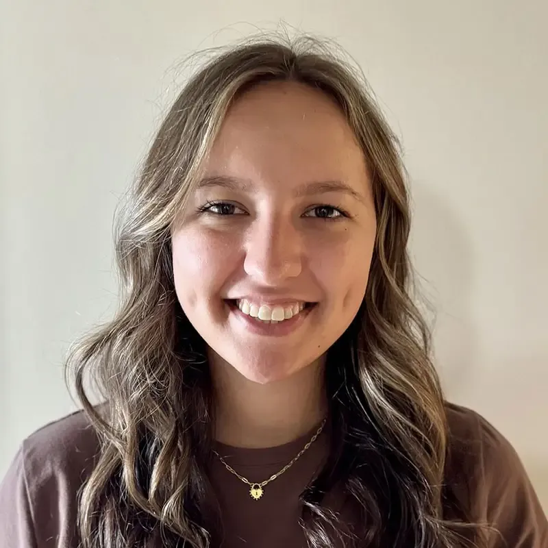 Person with long wavy brown hair smiling at the camera, wearing a brown top and a delicate gold necklace with a small pendant.