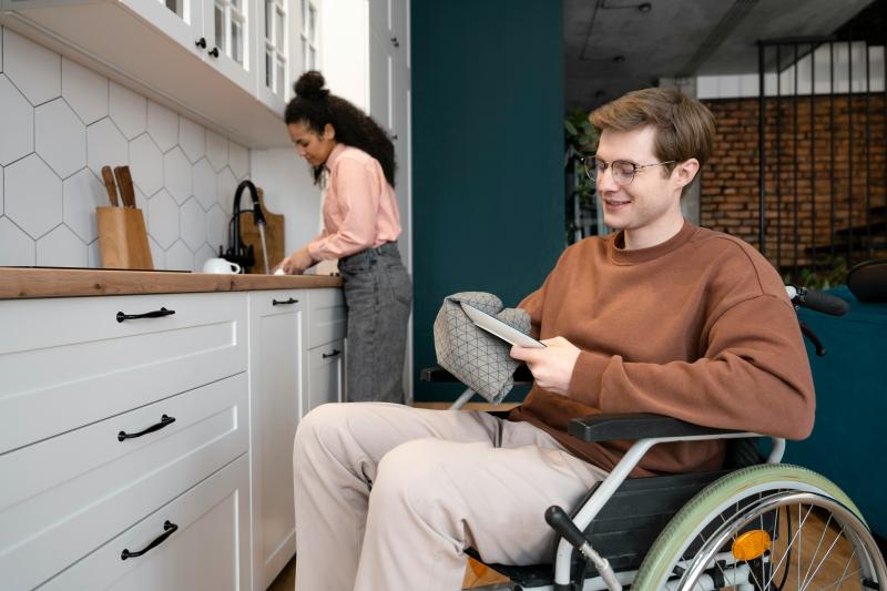 An individual is seen in the foreground in a wheelchair cleaning a dish, another individual assists in the background