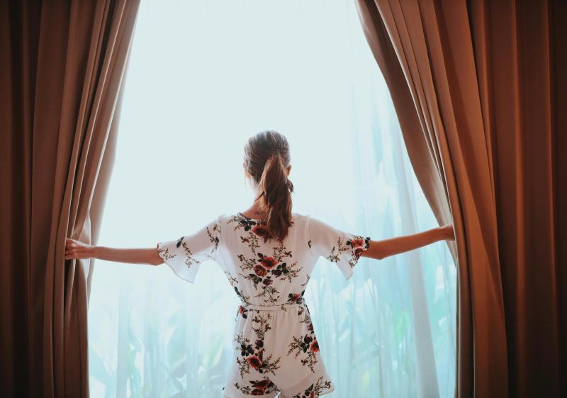 A person in a white floral dress and ponytail opens light orange curtains at a bright, sunny window.