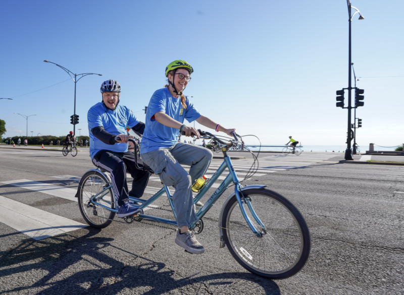 Two people ride a tandem bike along a lakeside path on a sunny day.