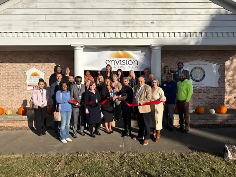 Group of community members and professionals cutting a red ribbon at the entrance of the Envision Unlimited mental health center building during its opening ceremony.