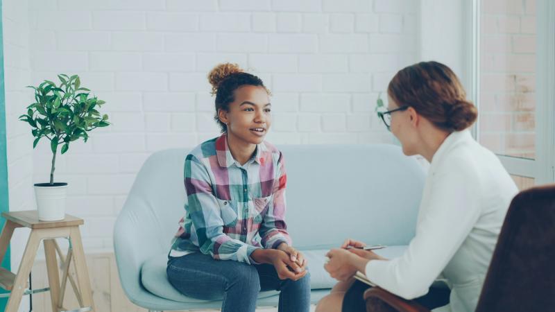 Two people have a conversation; one sits on a couch in a colorful plaid shirt and light blue pants while another in a white shirt and glasses sits in the foreground. A plant in a white pot rests on a wooden stool to the far left.