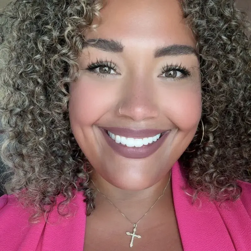 Person with curly dark hair smiling at the camera, wearing a bright pink top and a cross pendant necklace with silver hoop earrings.