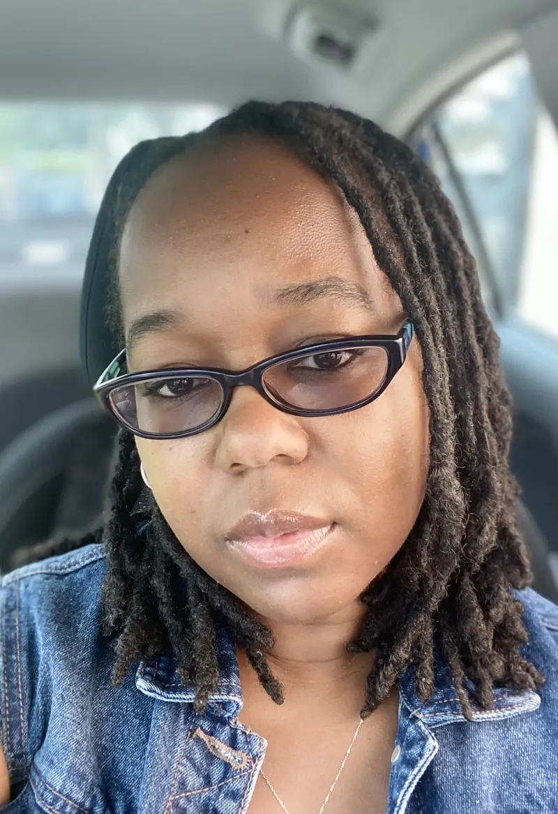 Close-up of a person with twists, wearing glasses and a denim jacket, sitting in a car.